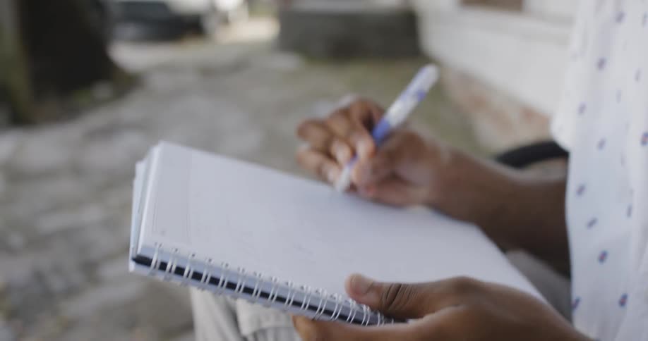 A young black man wearing glasses sits on a step outside of a building while he holds a composition notebook and is sketching out a picture with a pen