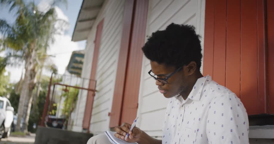 A young black man sits outside a house thinking and then writing in a notebook