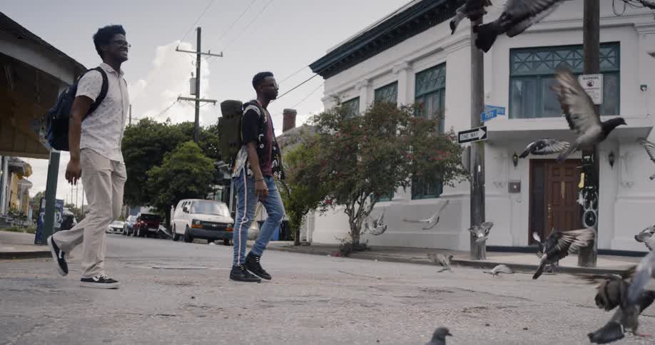 Two young men are wearing backpacks while crossing a city street filled with pigeons