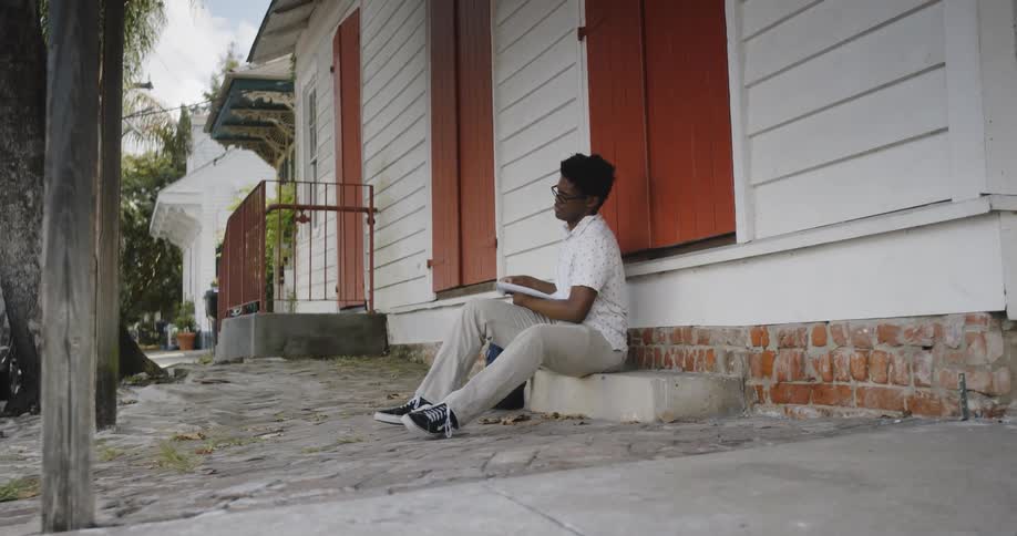 Young black man sits on step outside building writing in notebook