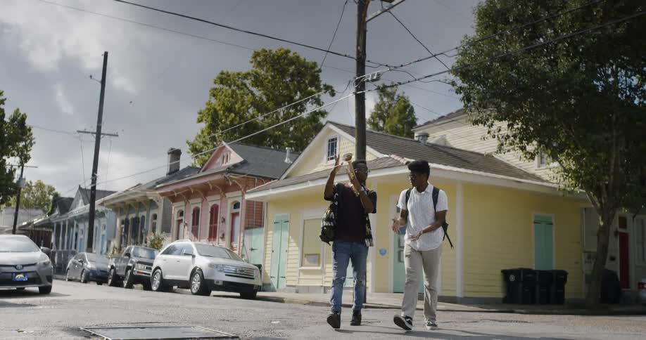 Two young black men cross the street laughing and talking one carries a musical instrument