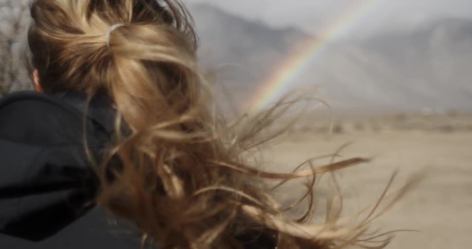 A blonde white woman stands in the desert and looks at a rainbow in the distance