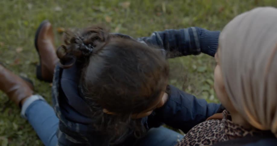 Woman wearing headscarf sits down and holds toddler to play on the grass in the daytime