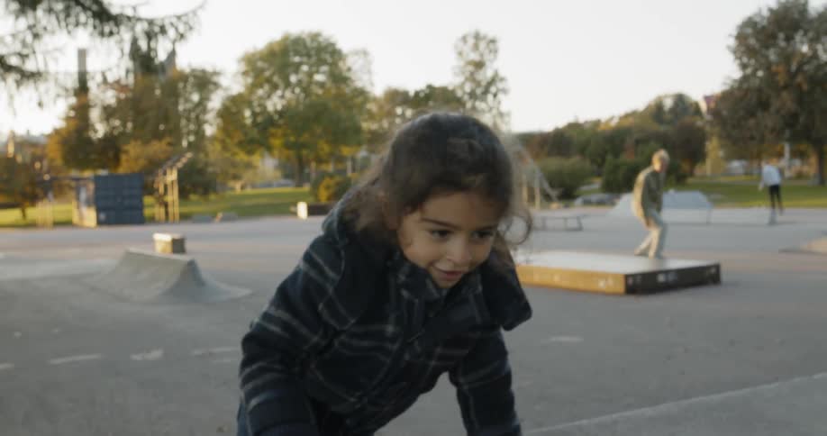 A young girl climbs and crawls on the concrete at a skate park while people skateboard in the background