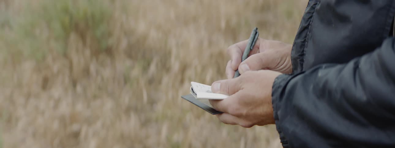 A white man wearing a hat and a coat writes something down in a small notebook