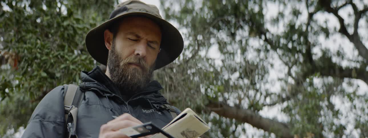 A man sketches in a book while standing under trees