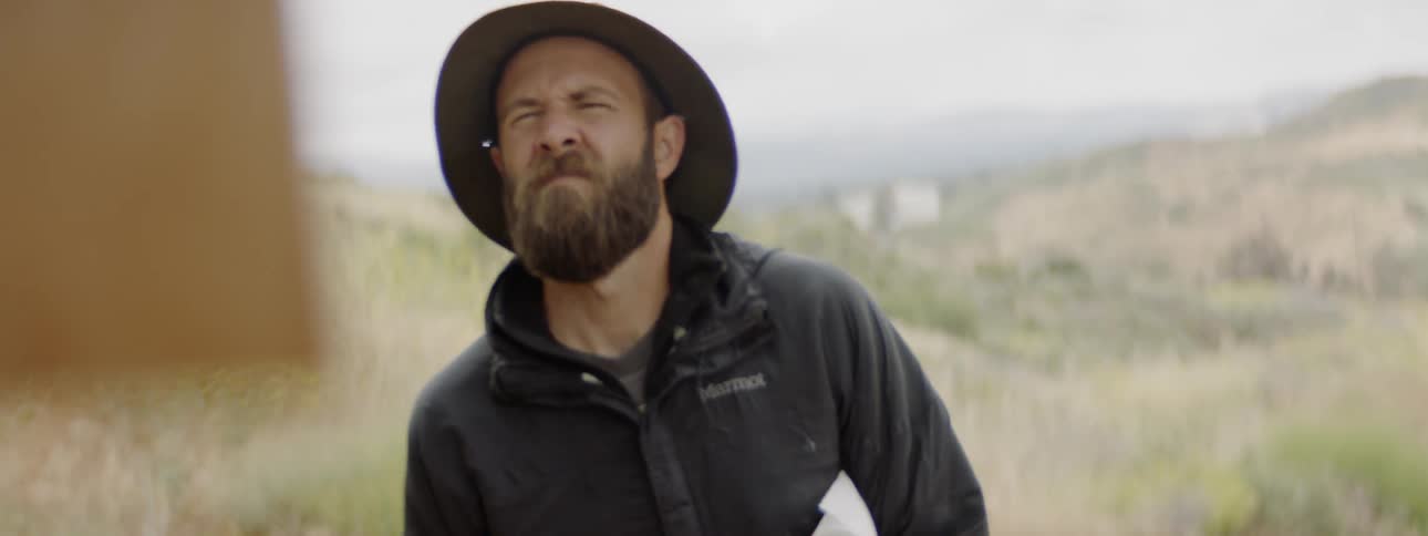 Bearded white man wearing hat looks at the frame on the hill outdoors