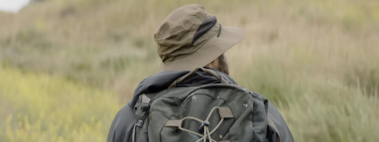 A white man with a hat and a backpack treks through grasslands
