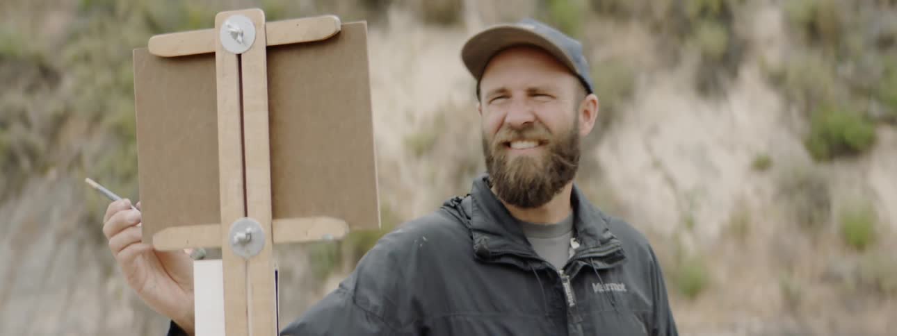 Bearded white man looks at the landscape and paints on the cardboard in the daytime