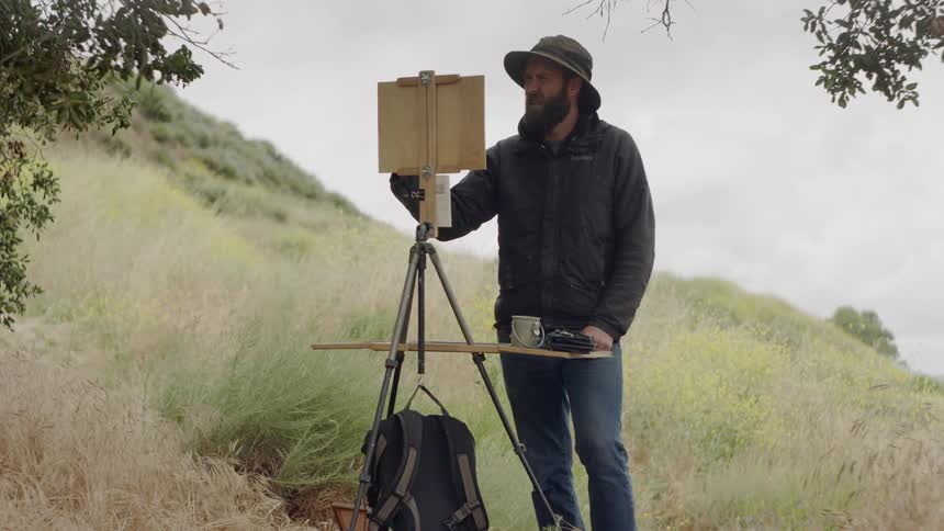 Man with beard steps back to look at painting on easel on hillside