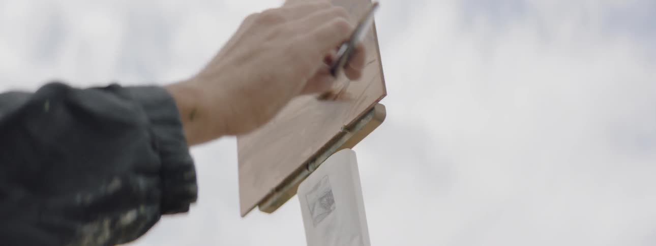A person's hand can been seen as they skillfully apply brushstrokes to a small painting as they stand outdoors