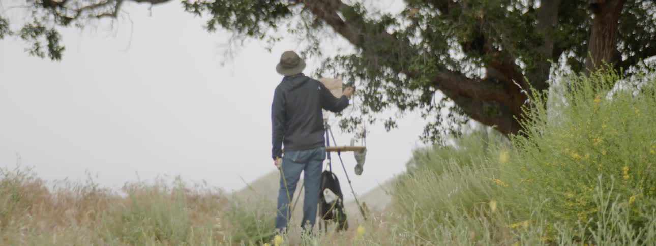 A man stands under a tree and paints on a small canvas