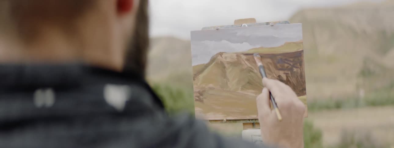 A white man wearing a hat paints a landscape picture outdoors