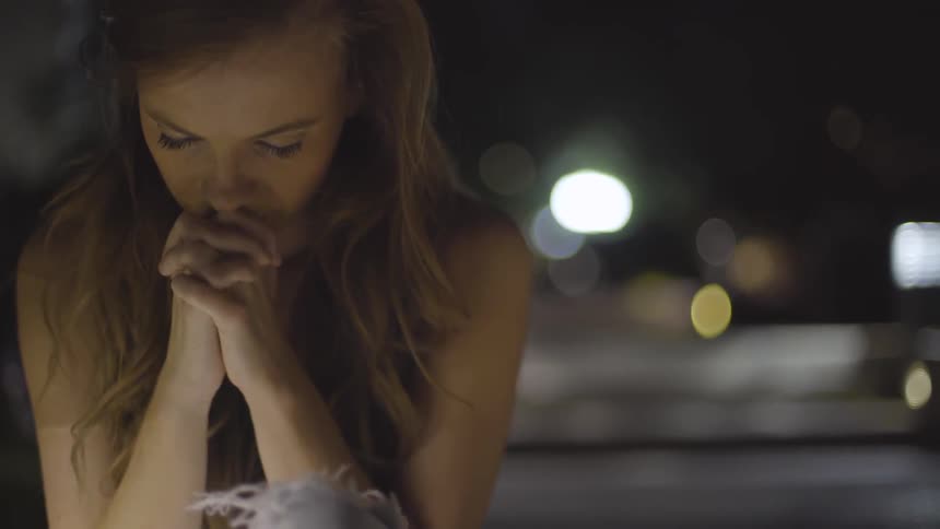 A woman stands in the dark looking down with her hands clasped together