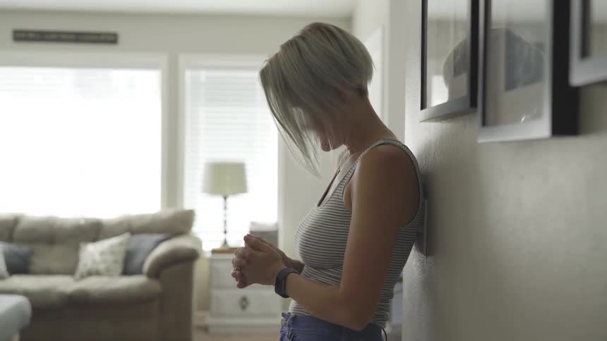A woman holds her hands as she leans up against a wall in the house at daytime