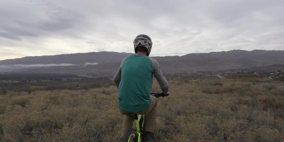 A man is riding a bike along a grass filled field on a cloudy day