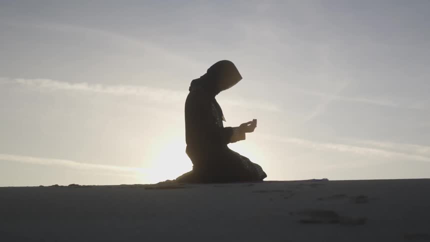 A person kneeling on the sand praying with the sun in the background