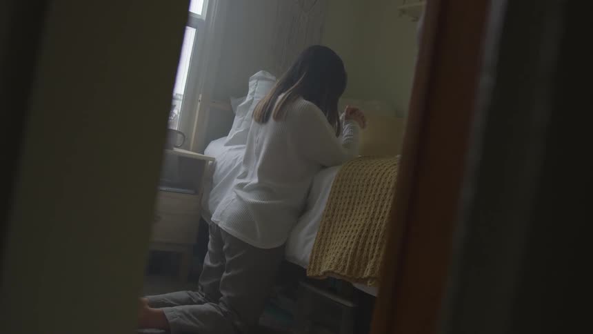 A young woman kneels by her bed praying