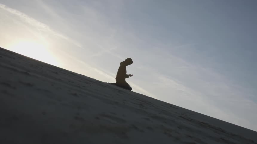 A woman kneels on a sandy slope and prays
