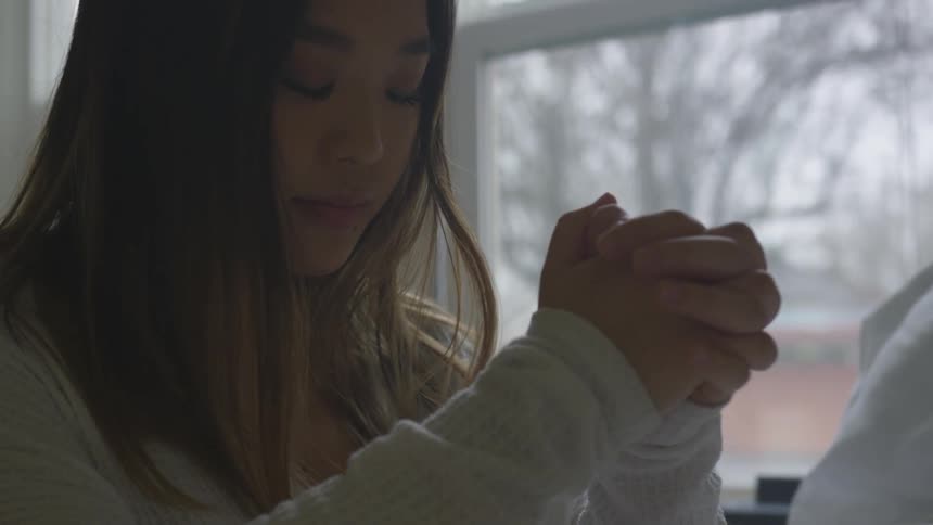 A female sitting down near a window with closed eyes and folded hands