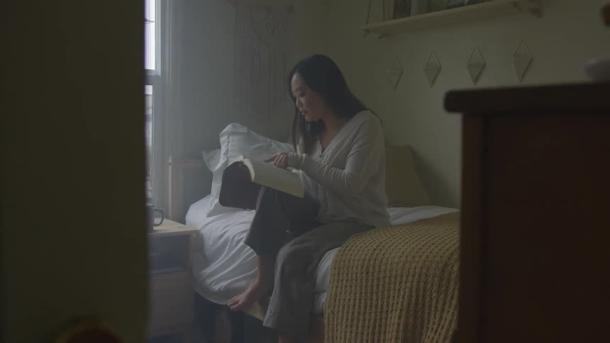 Woman sits on side of bed reading near window