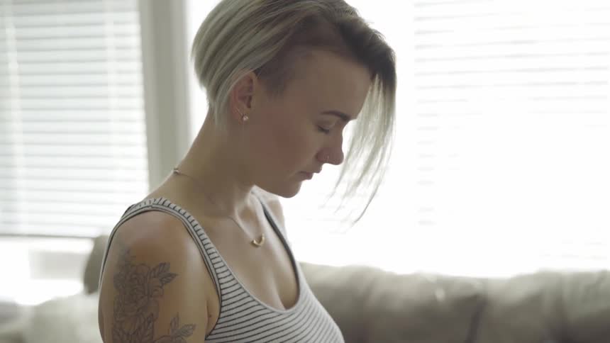 A woman with a tattoo on her arm stands with her head bowed and eyes closed next to a couch