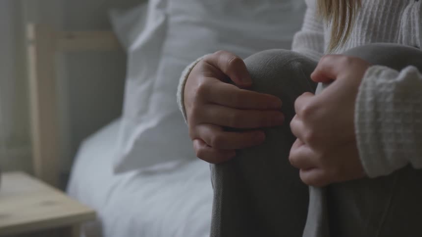 A woman holds her knees while sitting on a bed
