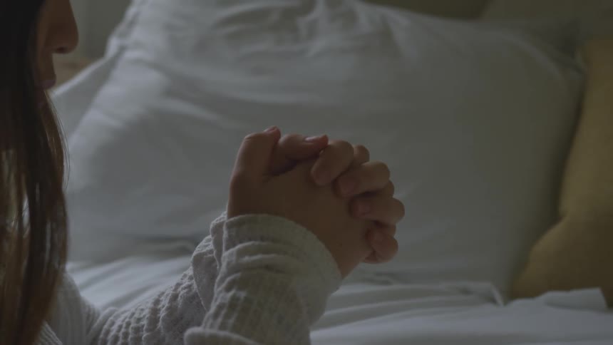 Woman prays with hands crossed at bedside in bedroom