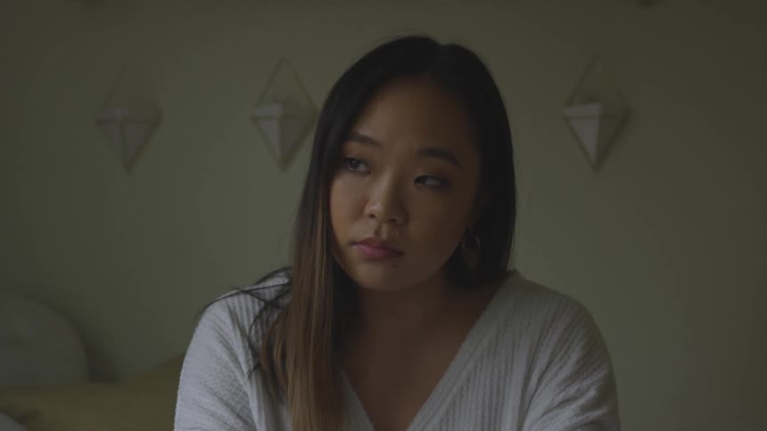 An asian woman sitting on a bed stares at something off screen