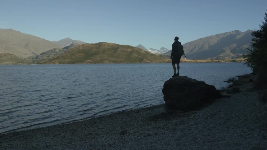 A man walks to the top of a rock and overlooks a large lake