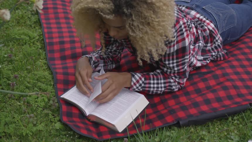 A woman is resting on a picnic blanket and reading a book