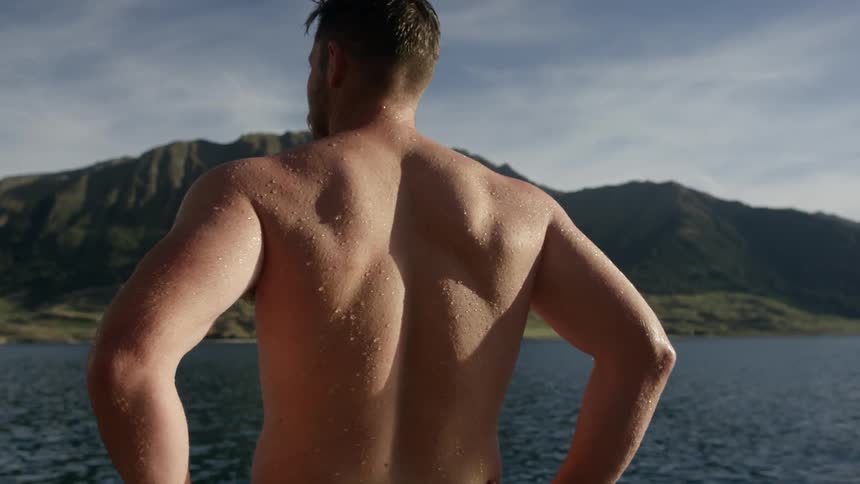 Wet and shirtless man stands in front of a lake
