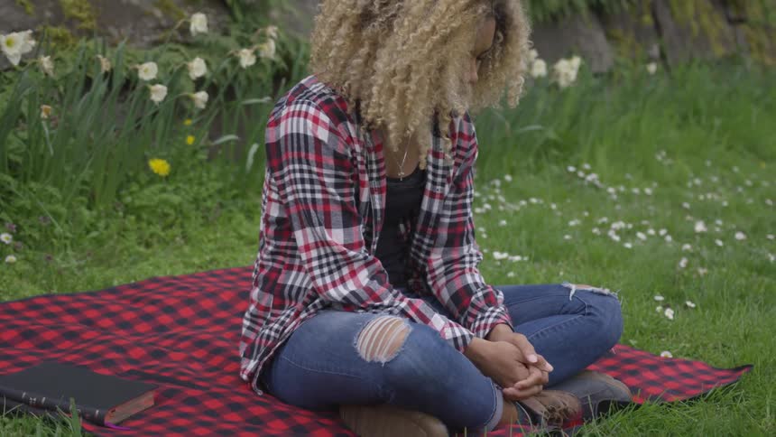 A woman with curly hair sits cross legged on a blanket in the grass