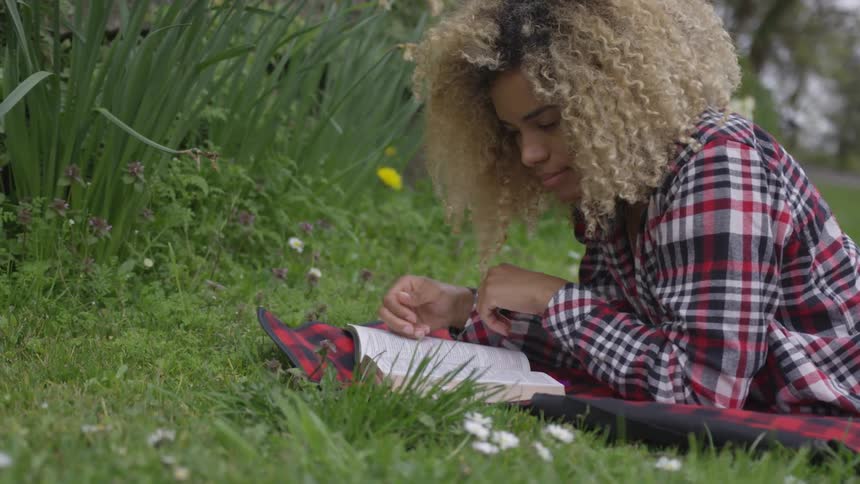 A black woman lies on a blanket on grass and reads a book