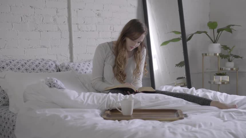 A woman is sitting on a bed and reading a book