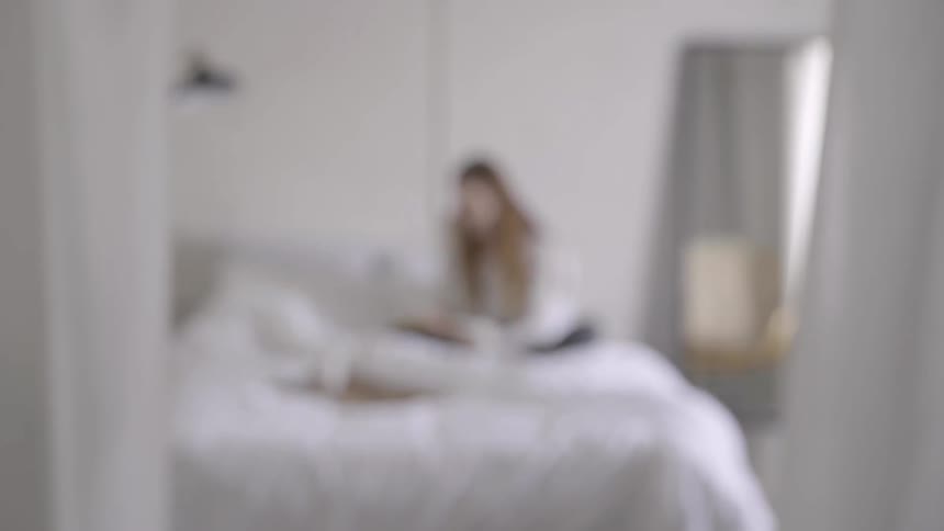 The long haired woman is sitting on the bed turning the pages of a book