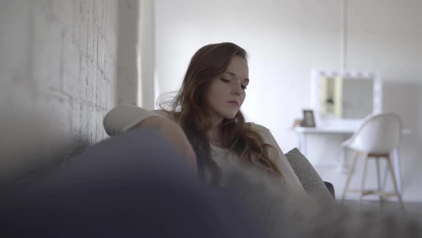 A white female looks deep in thought sitting on a sofa in a room with white walls and white furniture