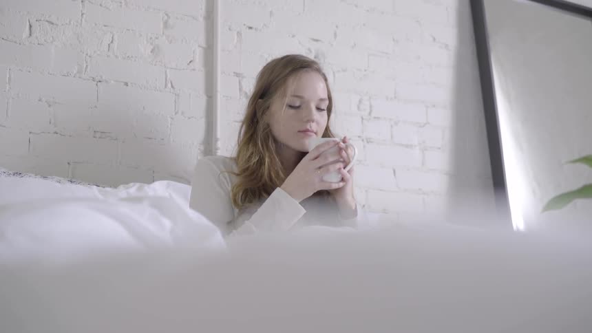 Young woman sitting on a bed smelling the contents of a mug