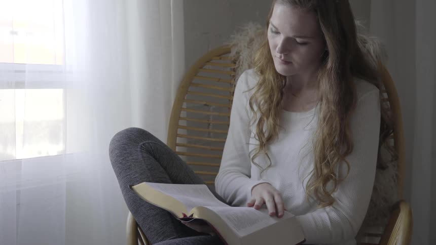 A woman that is sitting in a chair reads a book near a window