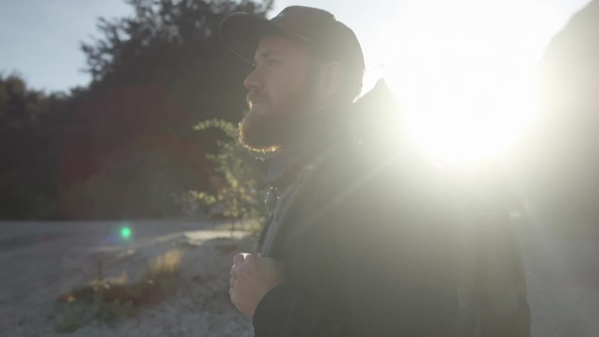 A bearded man carrying a backpack hikes along many trees on a sunny day