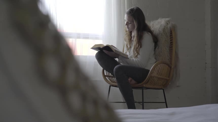 A woman sitting in a wicker chair reads a book by a window