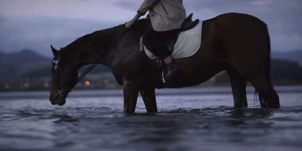 A young woman rides a horse through shallow water at dusk
