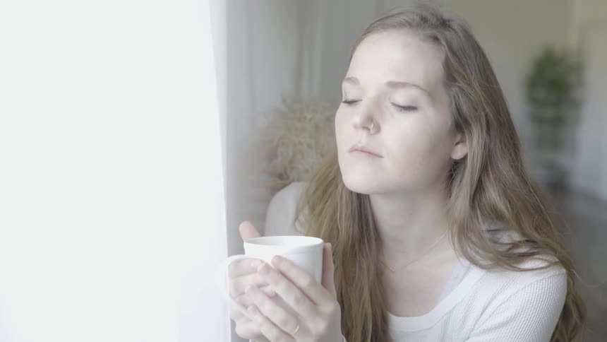 A young woman sits next to a window while holding a coffee mug