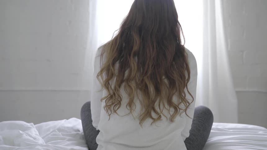 A woman with long brown hair sits on a bed and faces an open window with light entering the room