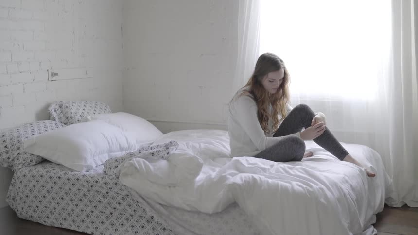 A young woman sits on a bed with her arms wrapped around her leg