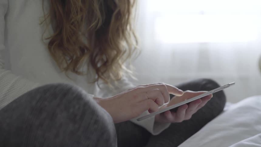 A woman with long hair sits cross legged on her bed scrolling on her tablet