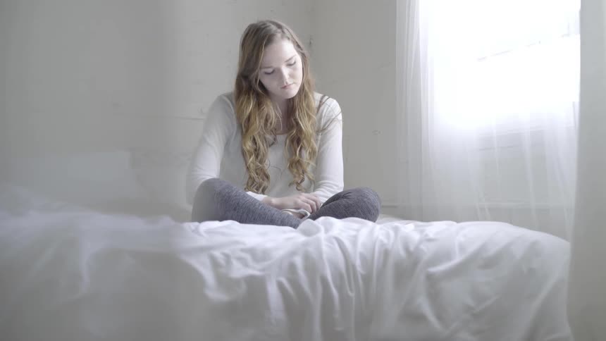 A woman sits on a bed and looks out a window
