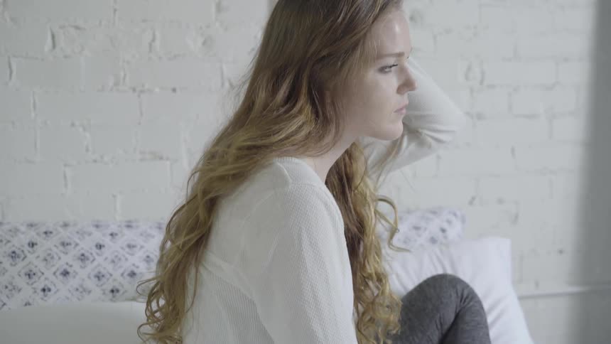 A sad looking woman runs her hand through her hair while sitting in bed