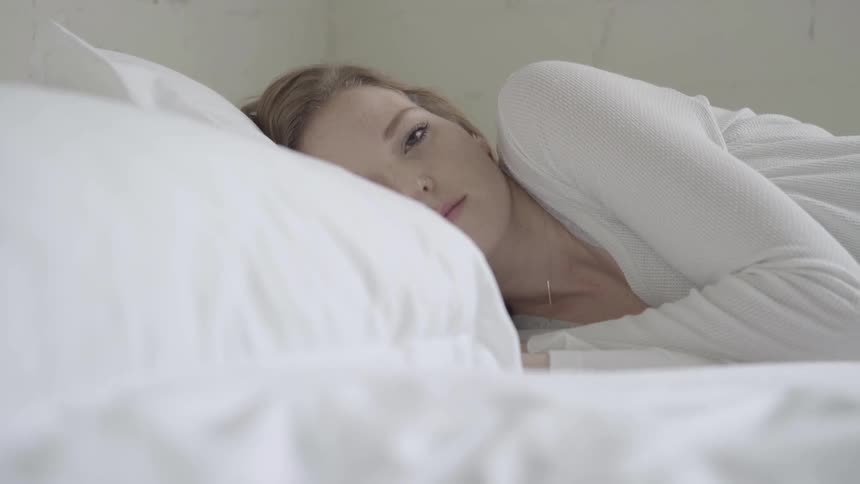 Woman wearing white clothes lies down on white sheets of the bed in the daytime