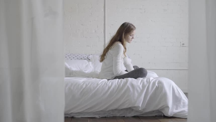 A sad looking woman sits on a bed with a white blanket